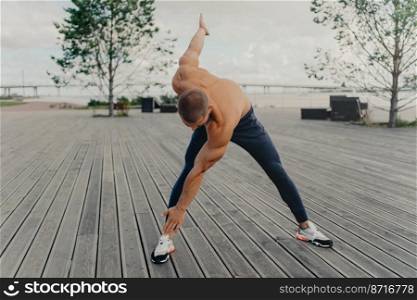 Handsome motivated unshaven sportsman does exercises on promenade, warms up before running, dressed in active wear, poses outdoor. Male runner with naked torso keeps fit, leads healthy lifestyle