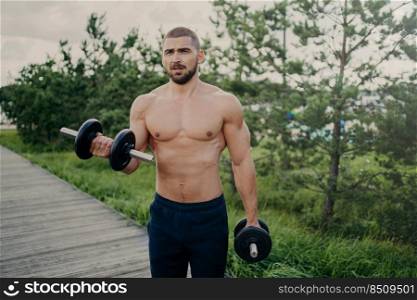 Handsome muscular man lifts barbells outside, does biceps training, stands with naked sexy torso, trains muscles, dressed in shorts poses outdoor, has attractive body. Weight lifting concept