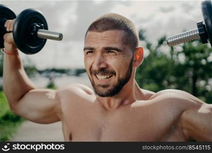Handsome muscular unshaven man with bristle raises dumbbells, does exercises and lifts barbells, stands with naked body outdoor. Strong bodybuilder makes weightlifting, poses with sport equipment