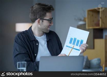 Handsome pleased German male entrepreneur in glasses representing outcome and income charts on laptop camera pointing with pencil during monthly online meeting while sitting at desk at home office. Smiling German male entrepreneur in glasses representing outcome and income charts on laptop camera