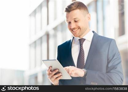 Handsome prosperous businessman reads useful information in internet via touchpad, connected to wireless internet, poses outdoor. Confident office worker makes booking online. Technology concept