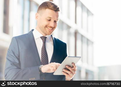 Handsome prosperous businessman reads useful information in internet via touchpad, connected to wireless internet, poses outdoor. Confident office worker makes booking online. Technology concept