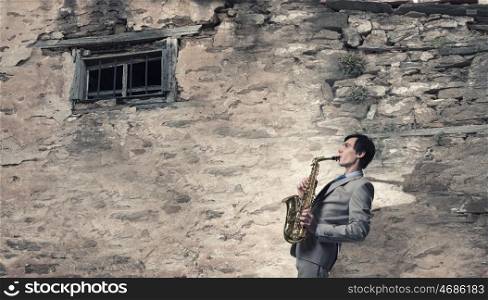Handsome saxophonist. Young man playing saxophone near window of old house