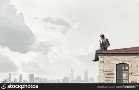 Handsome saxophonist. Young man sitting on roof and playing saxophone