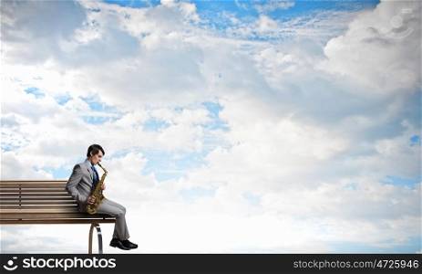 Handsome saxophonist. Young man sitting on wooden bench and playing saxophone