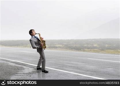 Handsome saxophonist. Young man walking on road playing saxophone