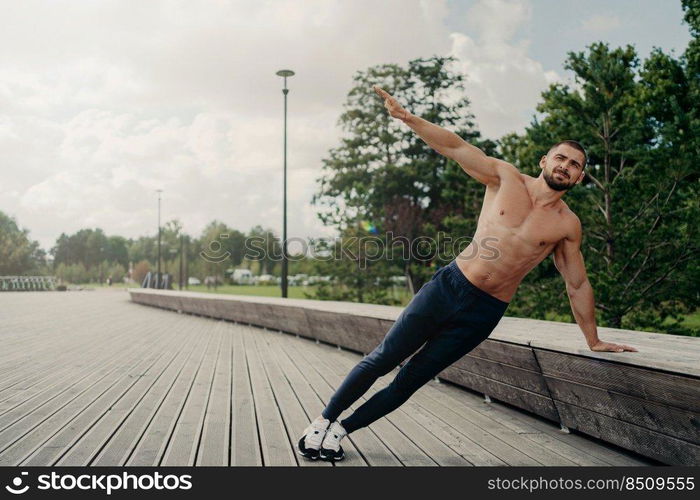 Handsome serious man stands in side plank, has perfect body constitution, poses with naked chest outdoors, keeps arm raised, trains for being healthy, dressed in sport trousers and sneakers.