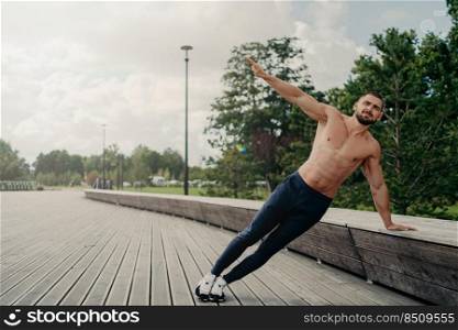 Handsome serious man stands in side plank, has perfect body constitution, poses with naked chest outdoors, keeps arm raised, trains for being healthy, dressed in sport trousers and sneakers.