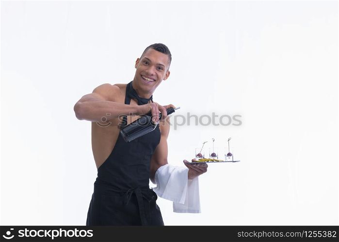 Handsome shirtless waiter holding a plate with glass shots and a bottle looks and smiles at the camera on a light background. Service and adult concept.. Shirtless waiter holding a plate smiles at the camera