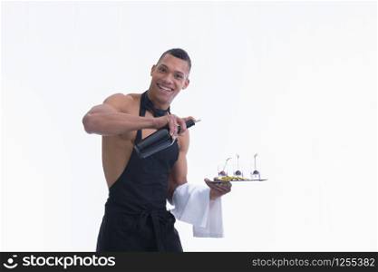 Handsome shirtless waiter holding a plate with glass shots and a bottle looks and smiles at the camera on a light background. Service and adult concept.. Shirtless waiter holding a plate smiles at the camera