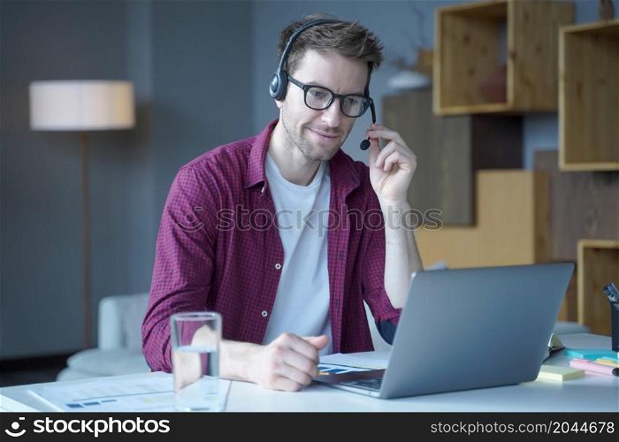 Handsome smiling austrian man in headset with microphone speaking by video call at home, friendly male call center agent consulting client online using laptop computer while sitting at his workplace. Friendly german guy call center agent consulting client online