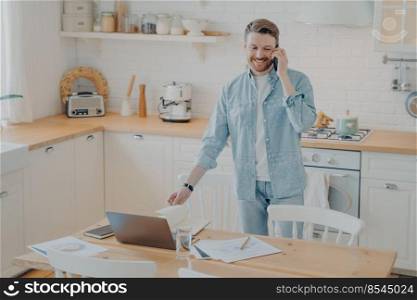 Handsome smiling bearded man wearing casual clothes working on laptop computer while standing at kitchen table at home and talking on mobile phone with friend. Freelance and distance work concept. Handsome smiling bearded man speaking on cellphone in kitchen while working remotely