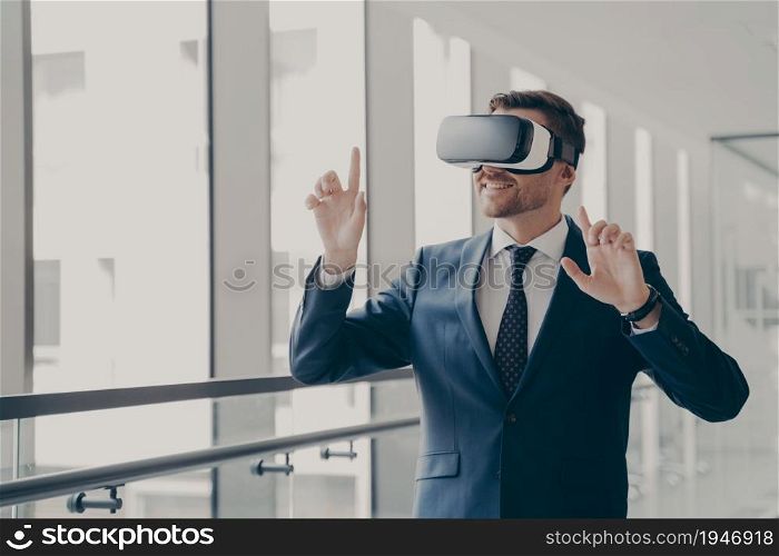Handsome smiling businessman in suit wearing vr headset standing in office and using innovative technologies for business, managing or editing project in virtual reality , office worker in 3d goggles. Handsome smiling businessman in vr headset using innovative technologies for business at work