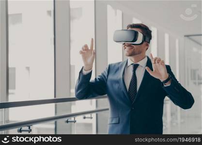 Handsome smiling businessman in suit wearing vr headset standing in office and using innovative technologies for business, managing or editing project in virtual reality , office worker in 3d goggles. Handsome smiling businessman in vr headset using innovative technologies for business at work