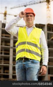 Handsome smiling engineer in hardhat posing against working construction crane