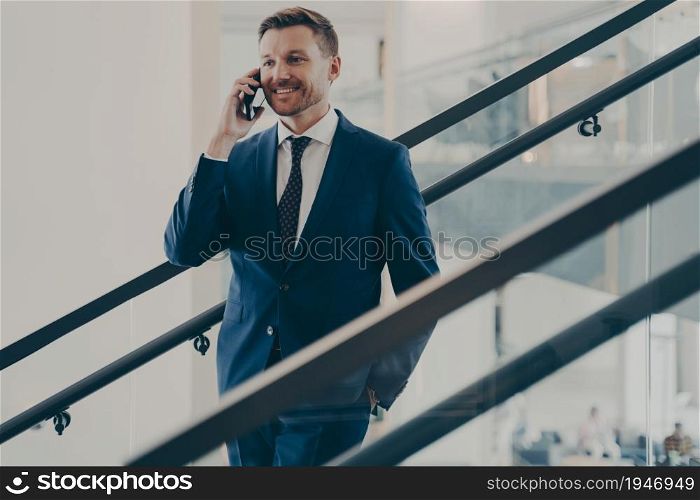 Handsome smiling european businessman in elegant blue suit having mobile conversation with client, making pleasant business call while standing on stairs in office, talking on smartphone at work. Handsome european businessman in elegant blue suit having mobile conversation in office