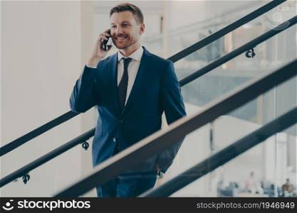 Handsome smiling european businessman in elegant blue suit having mobile conversation with client, making pleasant business call while standing on stairs in office, talking on smartphone at work. Handsome european businessman in elegant blue suit having mobile conversation in office