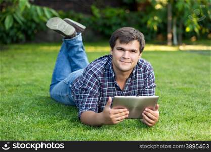 Handsome smiling student lying in park on meadow with digital tablet