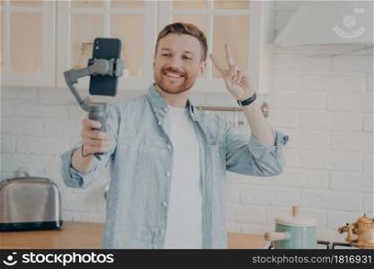 Handsome smiling young brunette man with stubble making selfie or streaming live on smartphone with gimbal stabilizer and gesturing peace sign in kitchen background at home, wears casual clothes. Handsome smiling young brunete man with stubble making selfie in kitchen