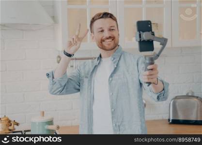 Handsome smiling young brunette man with stubble making selfie or streaming live on smartphone with gimbal stabilizer and gesturing peace sign in kitchen background at home, wears casual clothes. Handsome smiling young brunete man with stubble making selfie in kitchen