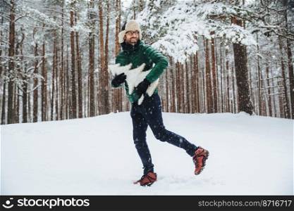 Handsome smiling young male wears warm hat and anorak, spends free time outdoor in forest during winter time prepares for holidays, holds fir tree. Bearded guy in eyewear admires winter landscapes