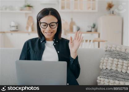 Handsome spanish girl in glasses has video call on laptop and waving hand. Woman says hello to business partner. Remote work on quarantine. Businesswoman is communicating with colleagues for project.. Handsome girl in glasses has video call and waving hand. Communicating with colleagues for project.