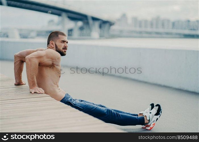 Handsome sportsman trains muscles and makes reverse push ups exercises warms up outdoors before run, concentrated aside, has muscular body, leads active healthy lifestyle, has bare muscular torso