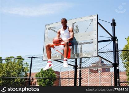 Handsome sporty African-American male basketball player dressed in white and holding his ball outdoor on a summer day in a basketball court while sitting in the hoop.