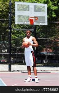 Handsome sporty African-American male basketball player with attitude dressed in white standing holding his ball with both hands outdoor on a summer day in a basketball court.