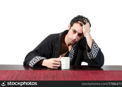 Handsome stressed guy in the morning who just woke up sitting at a table in his robe with a cup and hand in his hair, isolated on white