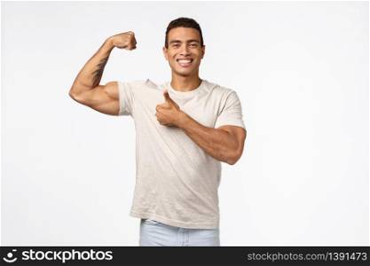 Handsome strong athletic sportsman in casual t-shirt, raise arm, tense muscle and proudly bragging good shape, big biceps, smiling pleased showing thumbs-up in approval and like, white background.. Handsome strong athletic sportsman in casual t-shirt, raise arm, tense muscle and proudly bragging good shape, big biceps, smiling pleased showing thumbs-up in approval and like, white background