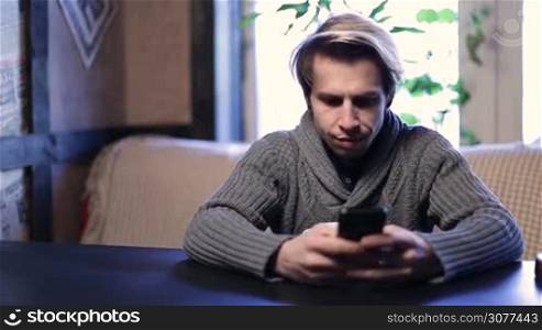 Handsome stylish hipster sitting at coffee shop and typing message on his mobile phone while drinking coffee. Smiling teenager using smart phone in cozy cafe. Cheerful young man spending leisure time at restaurant and texting on his cell phone.