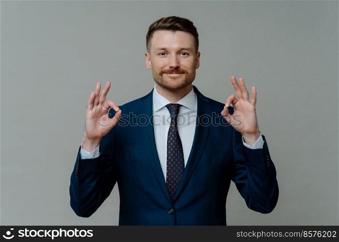 Handsome successful businessman in formal wear showing ok sign or okay gesture and smiling at camera while standing against grey background, male entrepreneur satisfied with done work. Happy businessman in suit showing that everything is ok