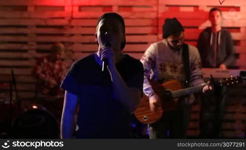 Handsome teenager vocalist holding microphone and singing with his band members playing musical instruments on background while band performing song in club. Rock group on stage at concert.