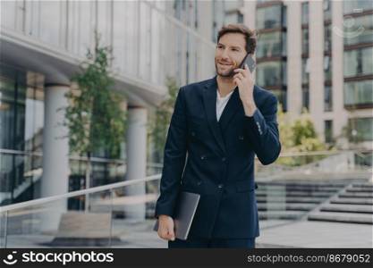 Handsome unshaven male entrepreneur communicates distantly with colleagues or partner uses modern technologies dressed in formal clothes poses outdoor near office building solves companies problems. Handsome unshaven male entrepreneur communicates distantly with colleagues or partner