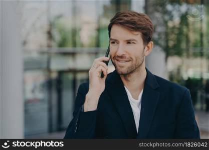 Handsome unshaven male executive worker left business building hears important information via speed mobile connectivity dressed in formal clothes poses against blurred background uses roaming tariffs. Handsome unshaven male executive worker left business building talks via smartphone