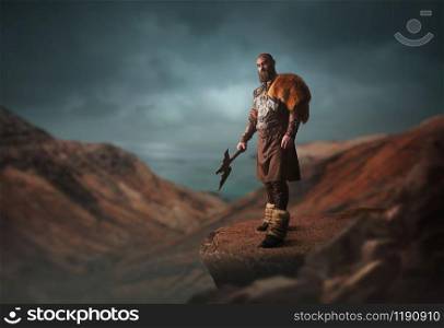 Handsome viking with axe dressed in traditional nordic clothes standing on the top of rocky mountain. Scandinavian ancient warrior