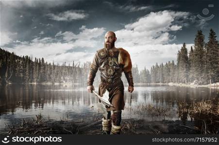 Handsome viking with axes in hands dressed in traditional nordic clothes standing at the lake. Ancient warrior at the river, northern forest on background