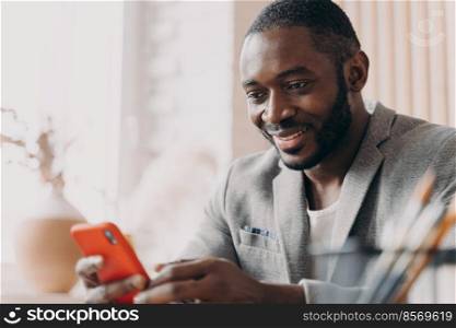 Handsome young african american businessman using mobile, chatting online while sitting at office desk, typing message and looking at phone screen with positive smile, distracted from work. Smiling african american businessman using mobile, chatting online while sitting at office desk