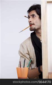 Handsome young artist with paintbrush in mouth contemplating