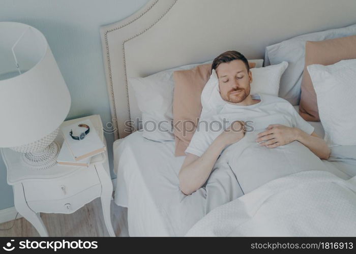 Handsome young bearded man peacefully sleeping in spacious comfortable bed in early morning at home, male enjoying sweet dreaming and relaxation during night sleep. View from above. Handsome young bearded man sleeping in spacious comfortable bed