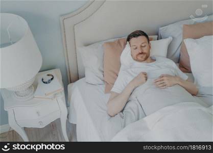 Handsome young bearded man peacefully sleeping in spacious comfortable bed in early morning at home, male enjoying sweet dreaming and relaxation during night sleep. View from above. Handsome young bearded man sleeping in spacious comfortable bed