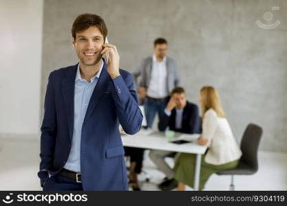 Handsome young business man standing confident in the office in front of his team and using mobile phone