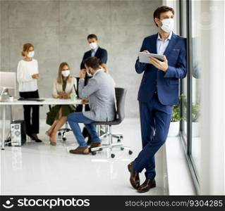Handsome young business man wearing protective facial mask while holding digital tablet in the office space