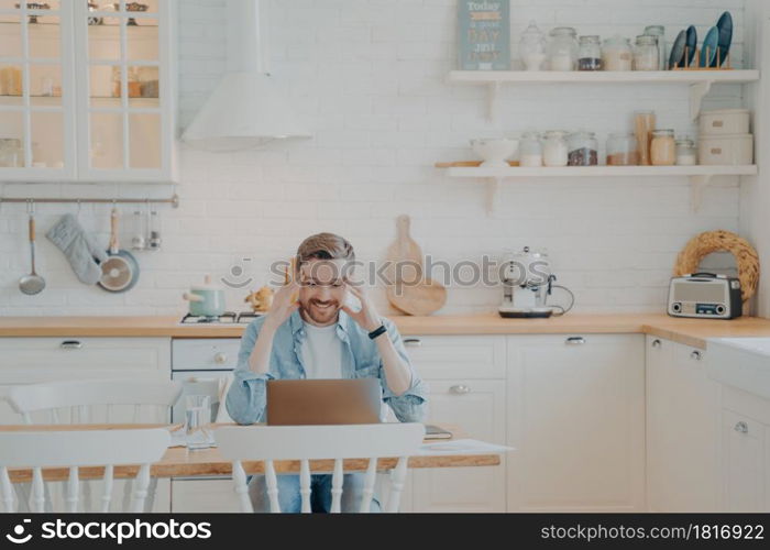 Handsome young businessman in casual clothes working with computer remotely while sitting at wooden table in stylish interior kitchen and smiling, looking at laptop screen with satisfied expression. Happy young businessman in casual clothes working with computer at home