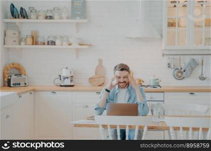 Handsome young businessman in casual clothes working with computer remotely while sitting at wooden table in stylish interior kitchen and smiling, looking at laptop screen with satisfied expression. Happy young businessman in casual clothes working with computer at home