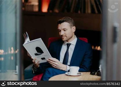 Handsome young businessman looks attentively at magazine, dressed in formal suit, drinks aromatic coffee or cappuccino, enjoys dinner break, calm armosphere in cozy restaurant. Business concept