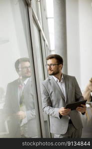 Handsome young businessman standing with digital tablet by the office window