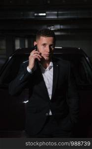 Handsome young businessman talking on the phone while standing near his car. A young man in a suit stands near the car and holds a telephone