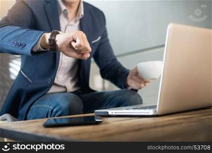 Handsome young businessman working at laptop with cup of coffee in restaurant. And looking at watch